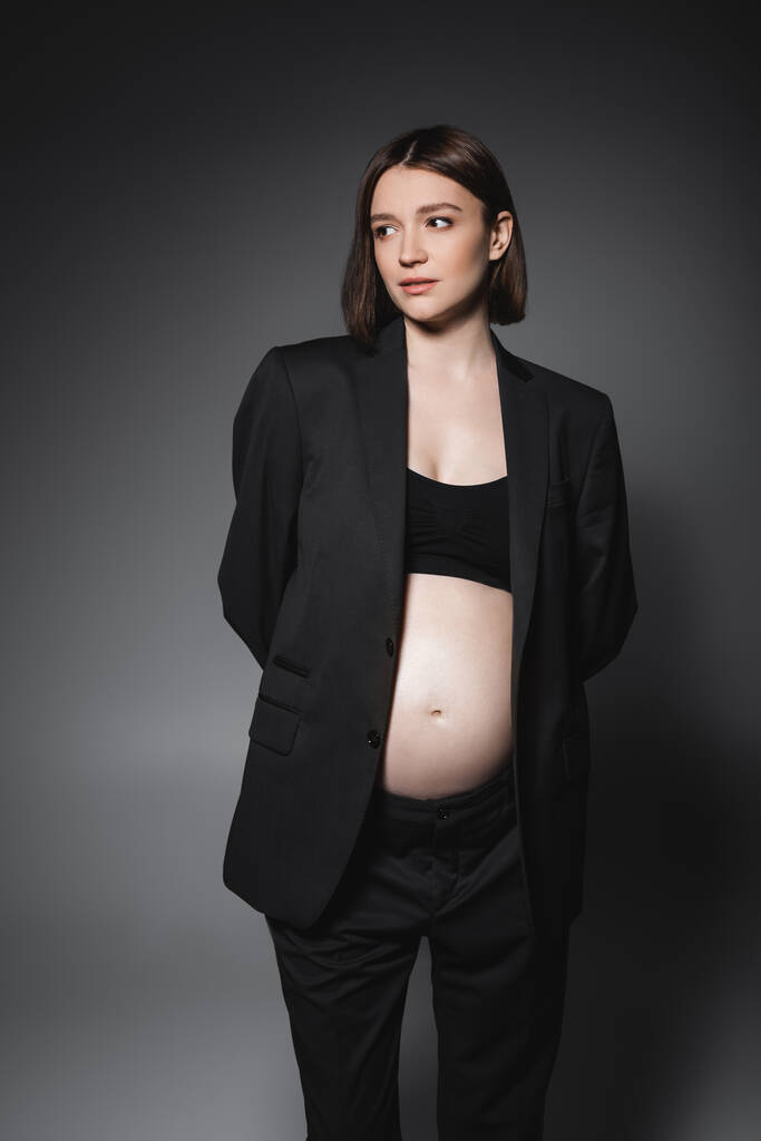 Νεαρή έγκυος γυναίκα με μαύρο μπλουζάκι και σακάκι κοιτάζει αλλού σε γκρι φόντο  - Φωτογραφία, εικόνα