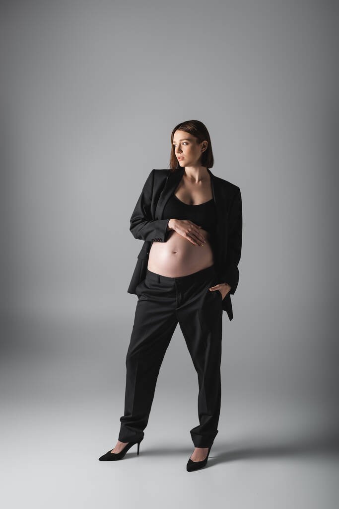 Hamile kadın takım elbiseli, göbeğine dokunuyor ve gri arka planda el ele tutuşuyor.  - Fotoğraf, Görsel