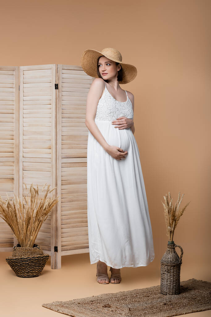Έγκυος γυναίκα με καπέλο ήλιο και φόρεμα αγγίζοντας την κοιλιά κοντά σε φυτά σε μπεζ  - Φωτογραφία, εικόνα