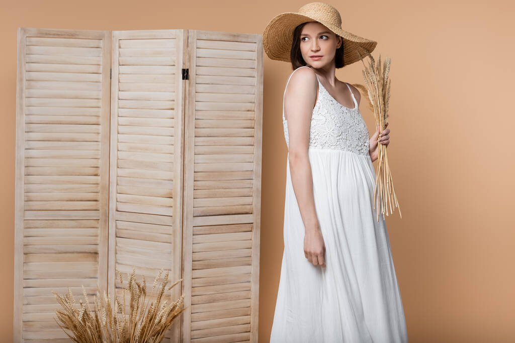 Trendy donna incinta in cappello di paglia in posa con spighette isolate sul beige  - Foto, immagini