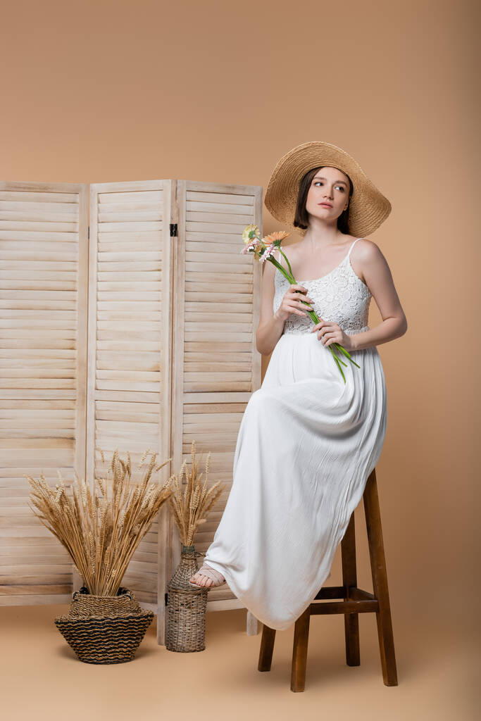 Elegante donna incinta in abito che tiene fiori vicino schermo pieghevole sul beige  - Foto, immagini