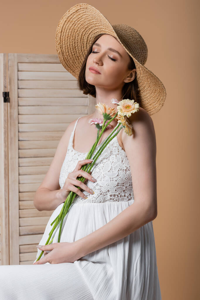 Έγκυος γυναίκα σε καπέλο ήλιο κρατώντας λουλούδια κοντά σε πτυσσόμενη οθόνη απομονώνονται σε μπεζ  - Φωτογραφία, εικόνα