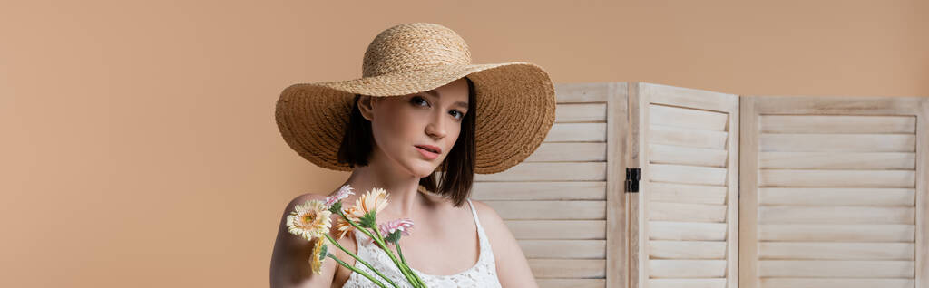 Brunette woman in straw hat looking at camera near flowers and folding screen isolated on beige, banner  - Photo, Image