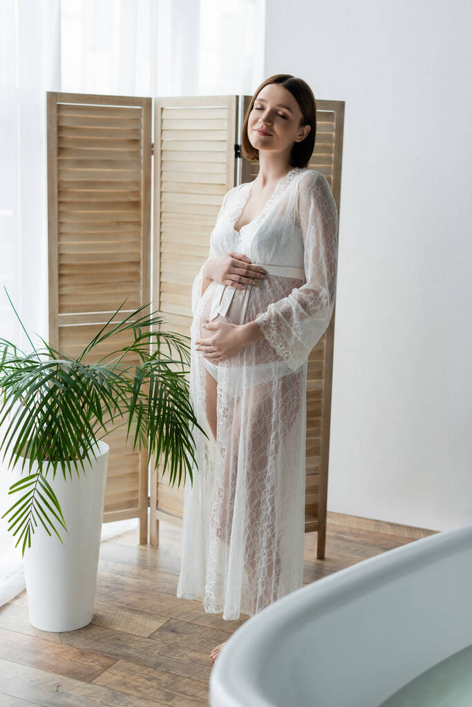 Удовлетворенная беременная женщина в халате стоит рядом с растением в ванной комнате  - Фото, изображение