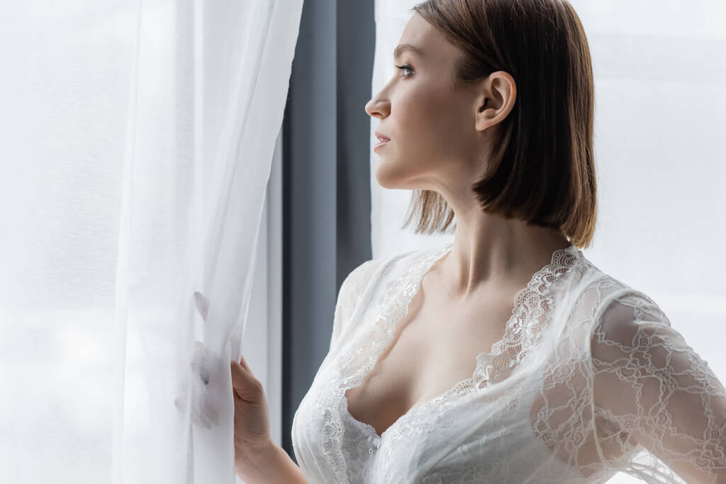 Vue latérale de la femme en robe de dentelle debout près de la fenêtre à la maison  - Photo, image