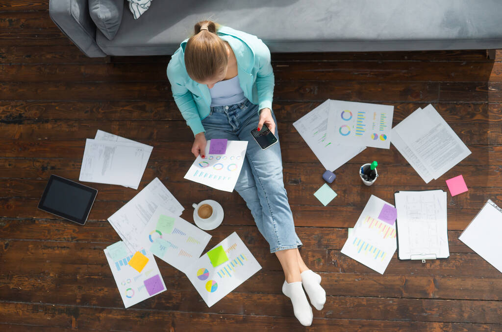 Mujer joven trabaja con documentos mientras está sentada en el suelo en casa. Estudiante, empresaria o freelancer que trabaja o estudia de forma remota a través de Internet. - Foto, imagen