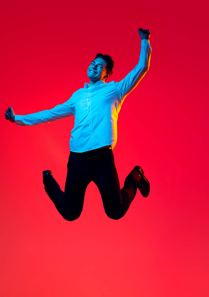 Retrato del joven hombre sonriente, estudiante de camisa blanca saltando alegremente excitado aislado sobre fondo rojo del estudio en neón azul - Foto, imagen