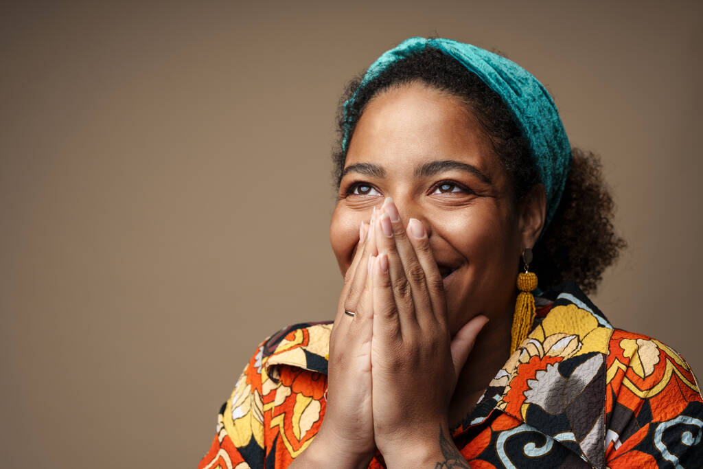 Mladá černošky žena v čelence s úsměvem a zakrývající ústa izolované přes šedé pozadí - Fotografie, Obrázek