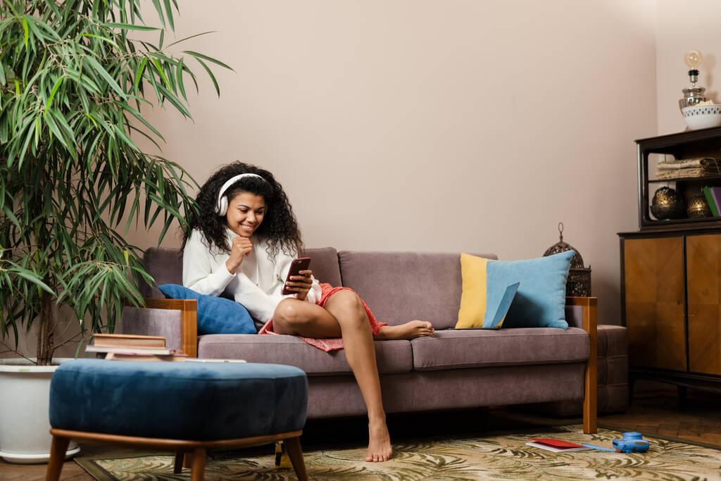Mladá černošky žena ve sluchátkách pomocí mobilu, zatímco sedí na pohovce doma - Fotografie, Obrázek