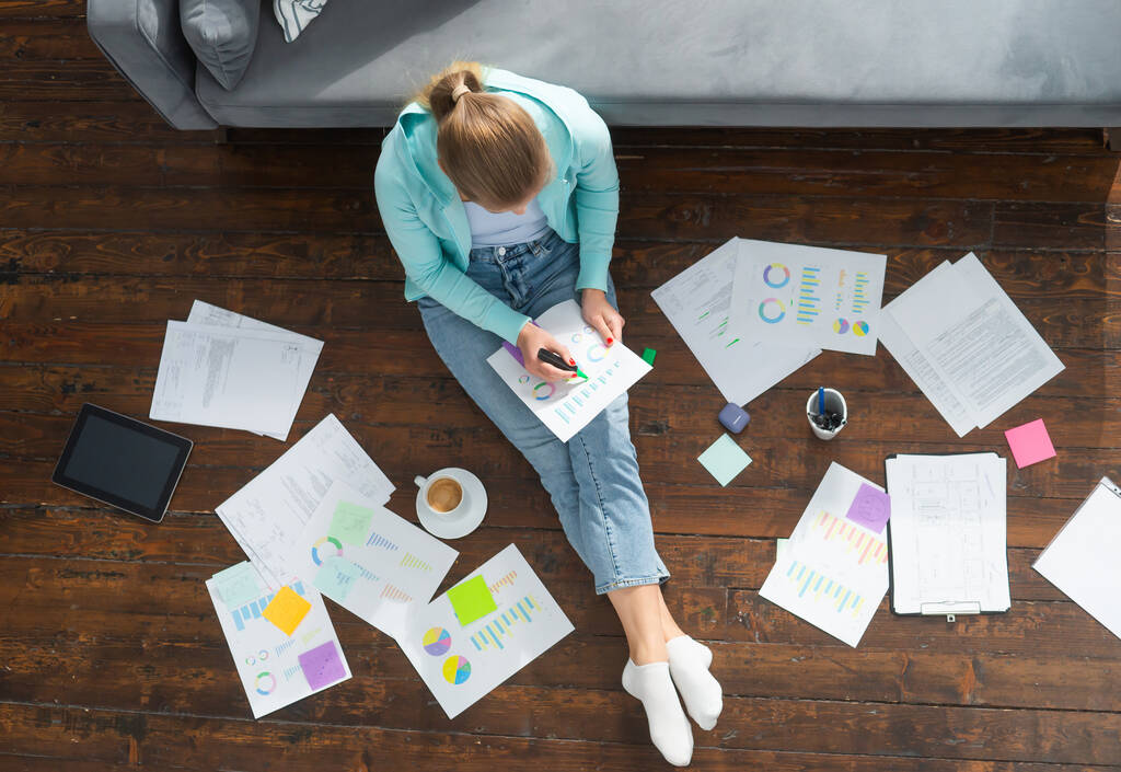 Молодая женщина работает с документами, сидя дома на полу. Студентка, предприниматель или девушка-фрилансер, работающая или обучающаяся дистанционно через интернет. - Фото, изображение
