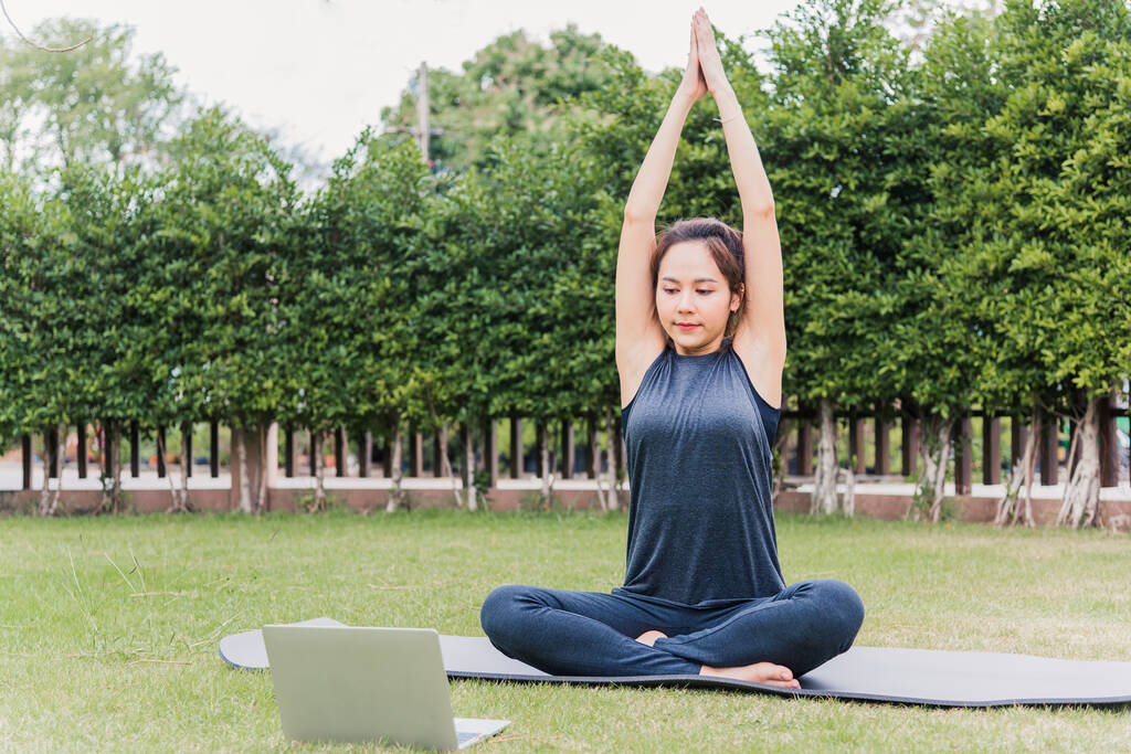 オンラインビデオトレーニング中にラップトップを使用してアジアの若い女性自然の中で緑の芝生の上に座って瞑想ポーズでヨガを屋外で練習フィールドガーデンパーク,ストレッチヘルスケアの概念 - 写真・画像