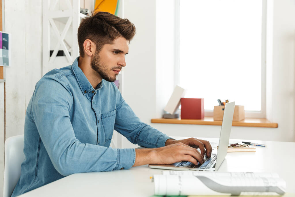 Jeune homme blanc travaillant avec un ordinateur portable assis au bureau à l'intérieur - Photo, image