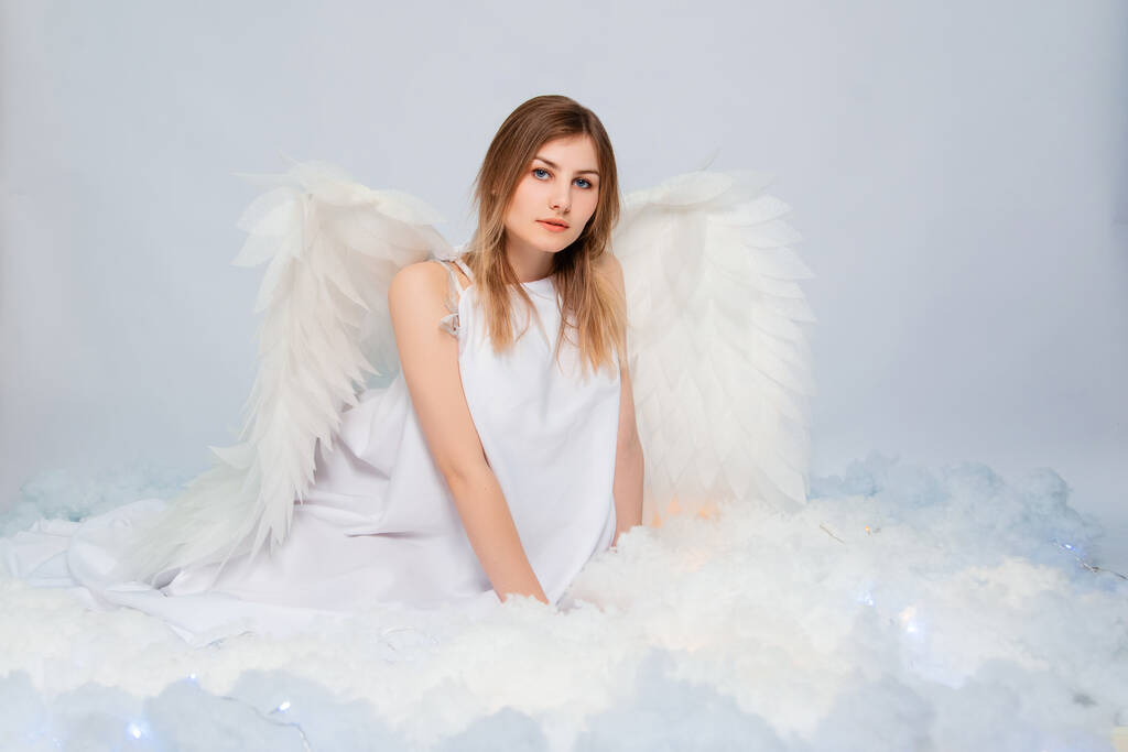 Una chica atractiva en una túnica blanca con grandes alas blancas detrás de su espalda posa mientras está sentada en nubes blancas y luminosas. - Foto, Imagen