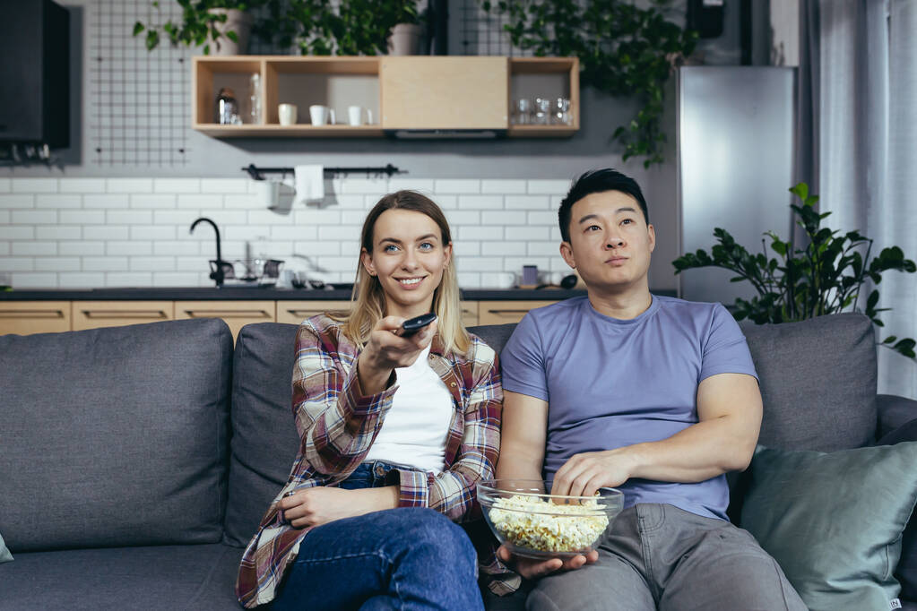 Jong stel in de liefde man en vrouw kijken tv samen thuis, verheugen en eten popcorn, gelukkig multiraciale familie in een nieuw appartement - Foto, afbeelding