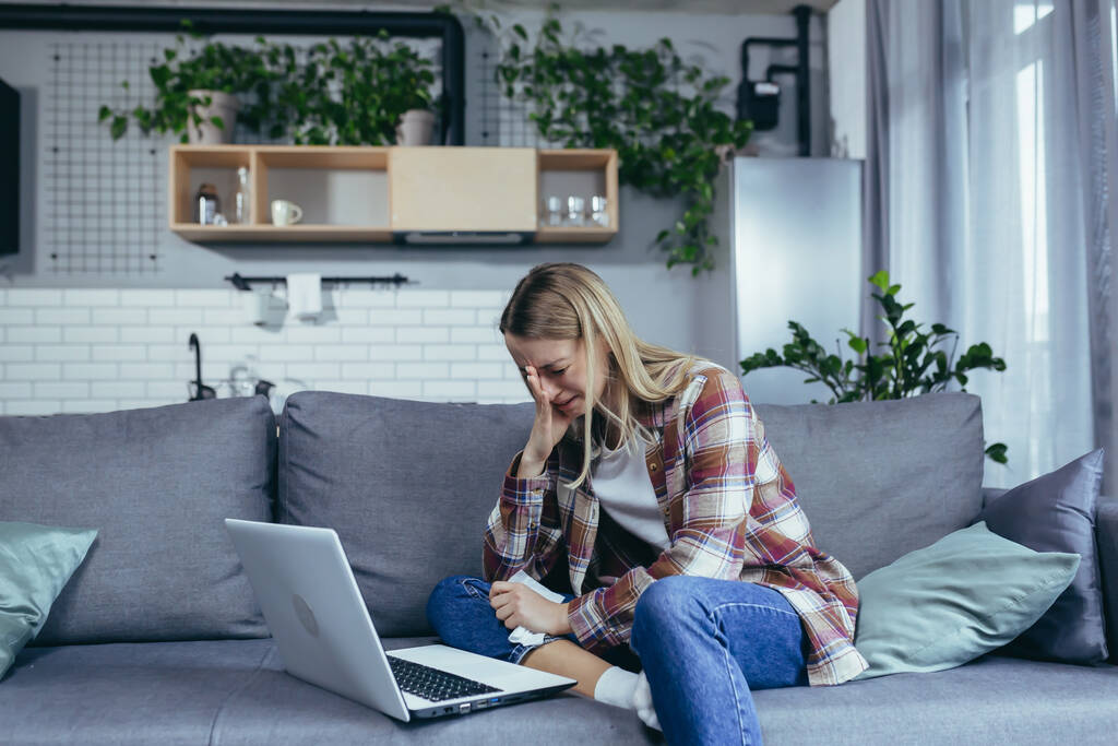 Junge schöne Frau allein zu Hause, nutzt Laptop für Online-Beratung mit einem Psychotherapeuten, telefoniert per Video mit der Hotline, setzt sich auf die Couch und weint - Foto, Bild