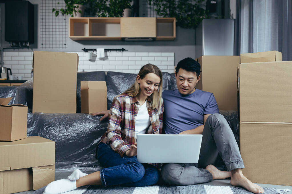 Liefdevol en gelukkig paar zitten op de vloer, Aziatische man en vrouw onder kartonnen dozen, kijken naar laptop scherm, online winkelen in online winkel - Foto, afbeelding