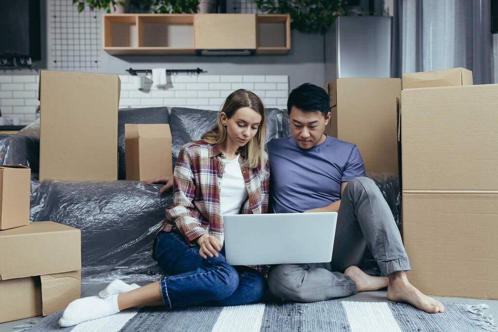 Молода сім'я в новій квартирі, продумано і серйозно вибирає нові меблі в інтернет-магазині, використовує ноутбук, сидячи на підлозі серед картонних коробок
 - Фото, зображення
