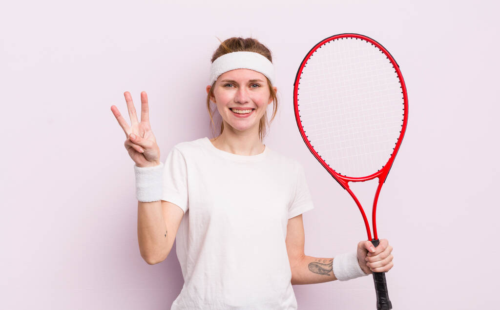 κοκκινομάλλα όμορφη κοπέλα χαμογελάει και φαίνεται φιλική, δείχνοντας νούμερο τρία. έννοια τένις - Φωτογραφία, εικόνα