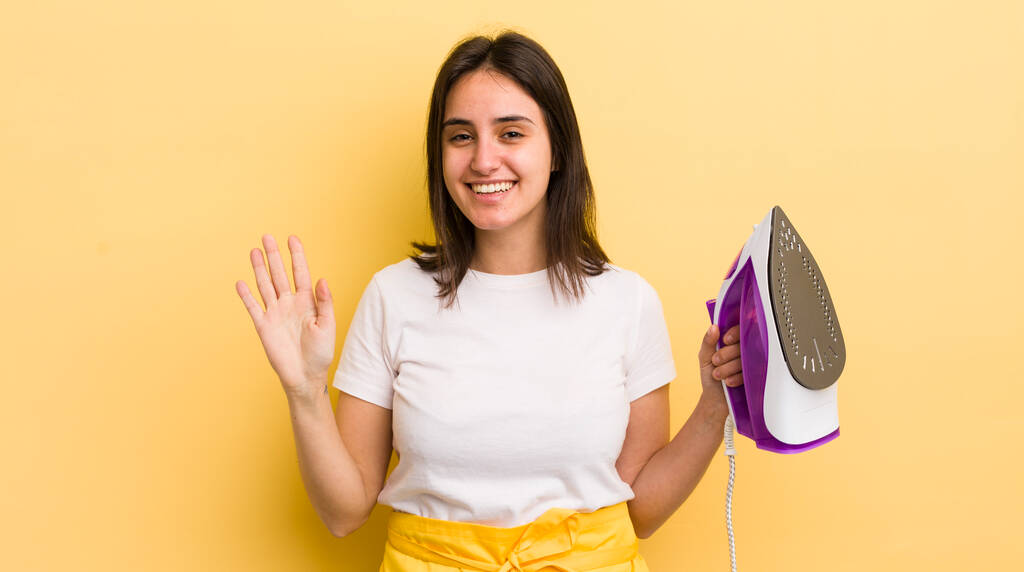 jonge Spaanse vrouw glimlachte vrolijk, zwaaide met de hand, verwelkomde en groette u. kleding ijzer concept - Foto, afbeelding