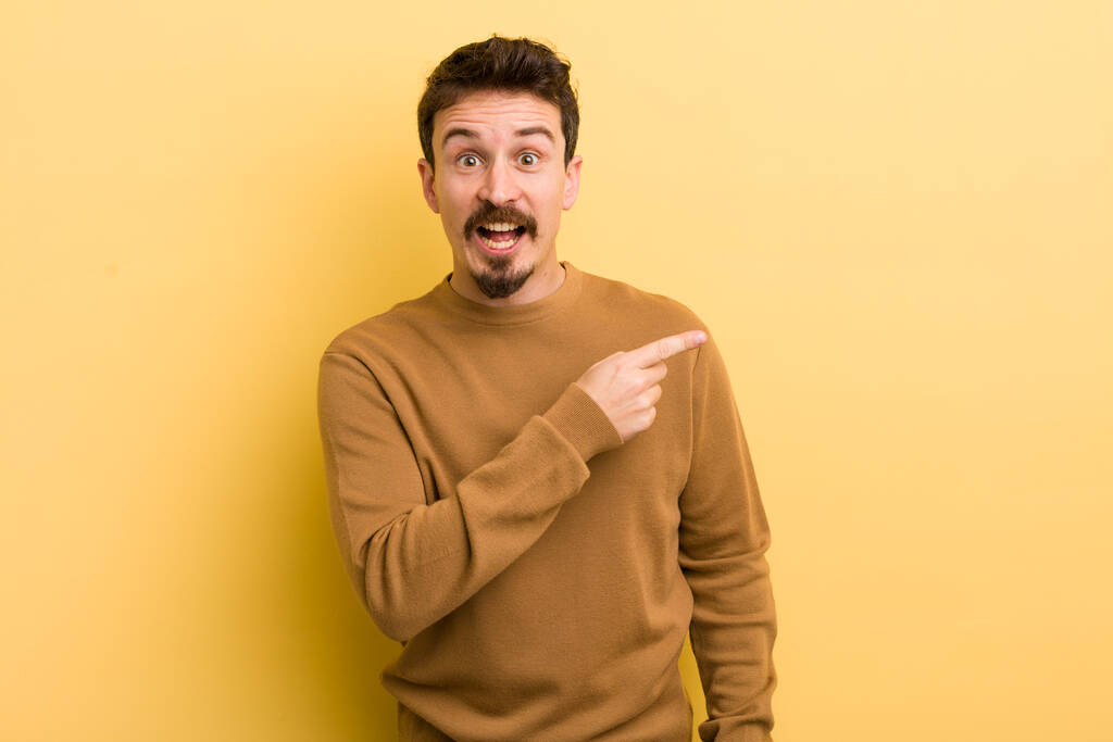 young hispanic man looking excited and surprised pointing to the side and upwards to copy space - Photo, Image