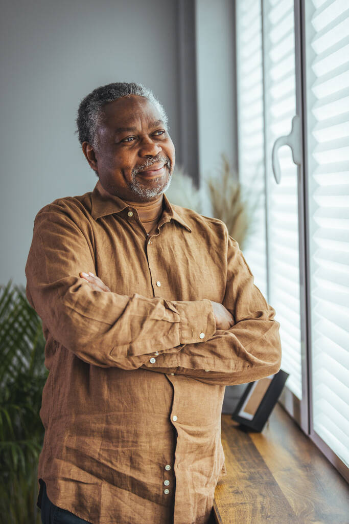 Вид сбоку на счастливого пожилого африканца в отставке, стоящего у окна дома. Удовлетворенный старик смотрит в окно и улыбается, стоя у окна. Положительные и уверенные в себе пожилые люди наслаждаются пенсией. - Фото, изображение