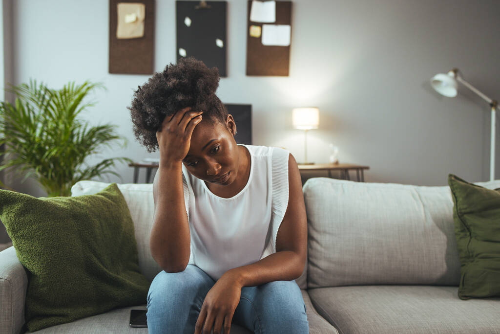 Mladá žena v šedých šatech sedí schoulená doma v posteli. Naštvaná Afroameričanka trpí a pláče. Deprese kvůli osamělosti, stresu, problémům. Pojem psychických poruch. - Fotografie, Obrázek