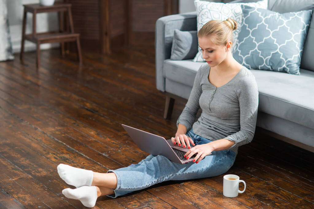 Młoda kobieta pracuje na laptopie siedząc w domu na podłodze. Student, przedsiębiorca lub freelancer dziewczyna pracuje lub uczy się zdalnie przez internet. - Zdjęcie, obraz