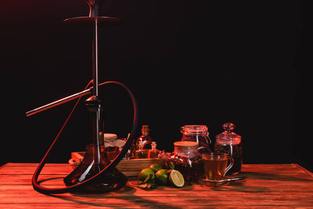 Té, limas frescas y narguile en mesa de madera aislada en negro con luz roja - Foto, imagen
