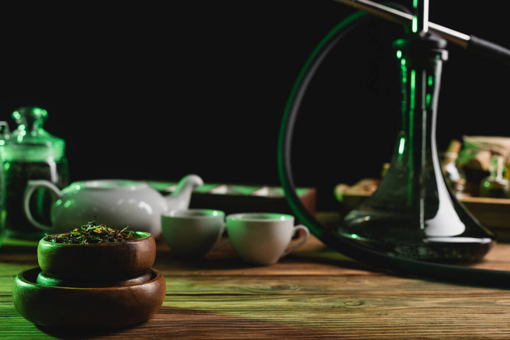 Сухой чай возле размытого кальяна и чашки на деревянном столе изолированы на черный  - Фото, изображение