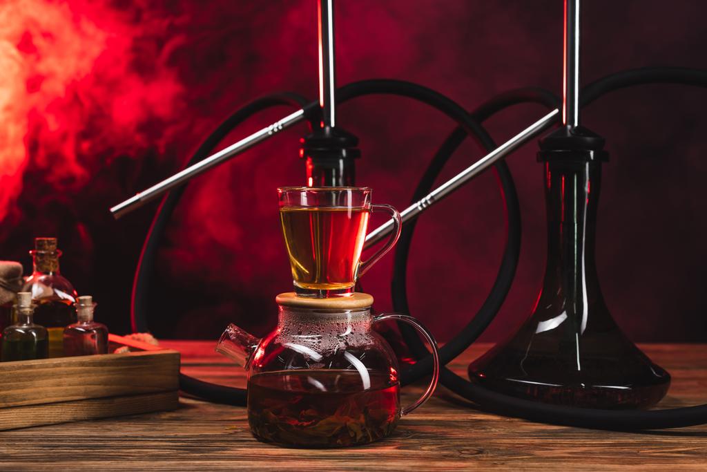 Taza de té en maceta cerca de hookahs en superficie de madera sobre fondo negro con humo rojo - Foto, Imagen
