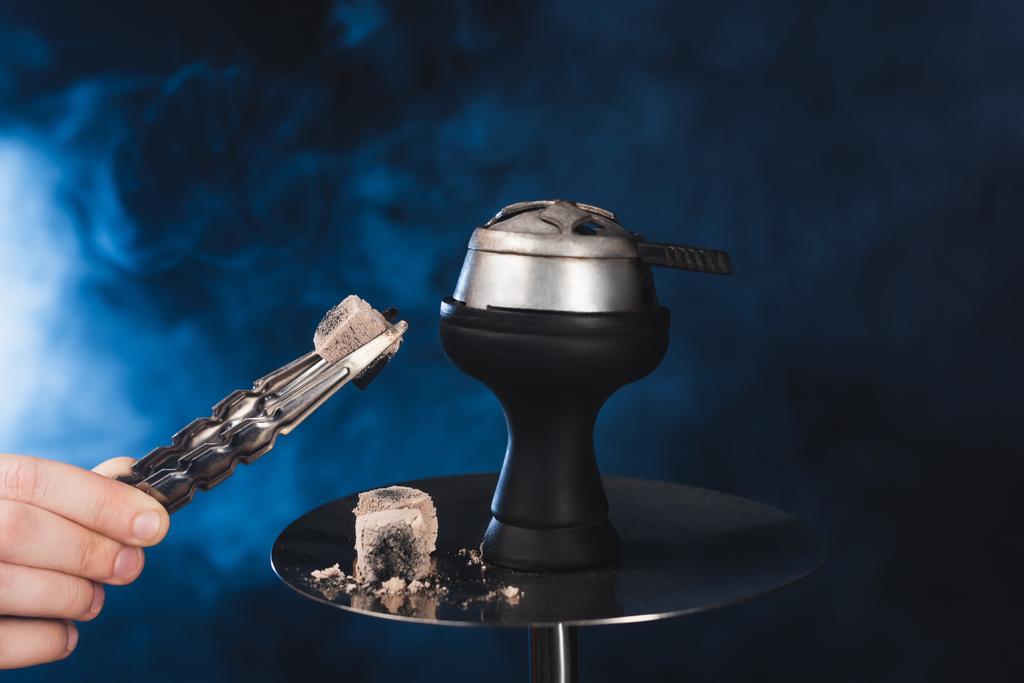 Cropped view of man holding tweezers with coal near hookah on black background with smoke - Photo, Image