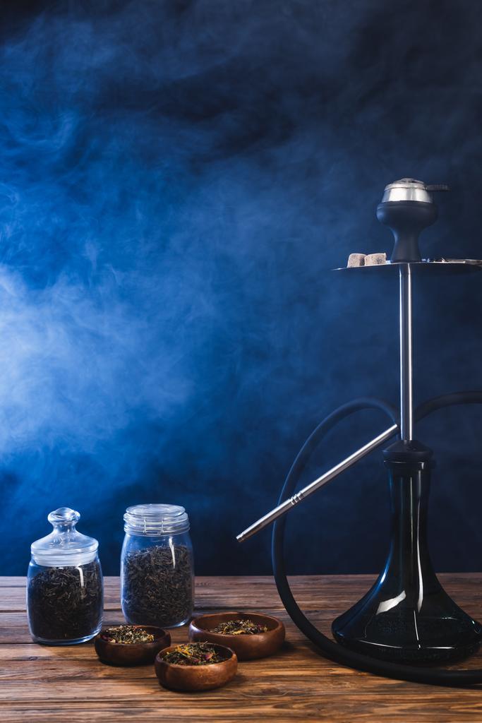 Кальян біля сухого чаю в мисках на дерев'яній поверхні на чорному тлі з синім димом
 - Фото, зображення