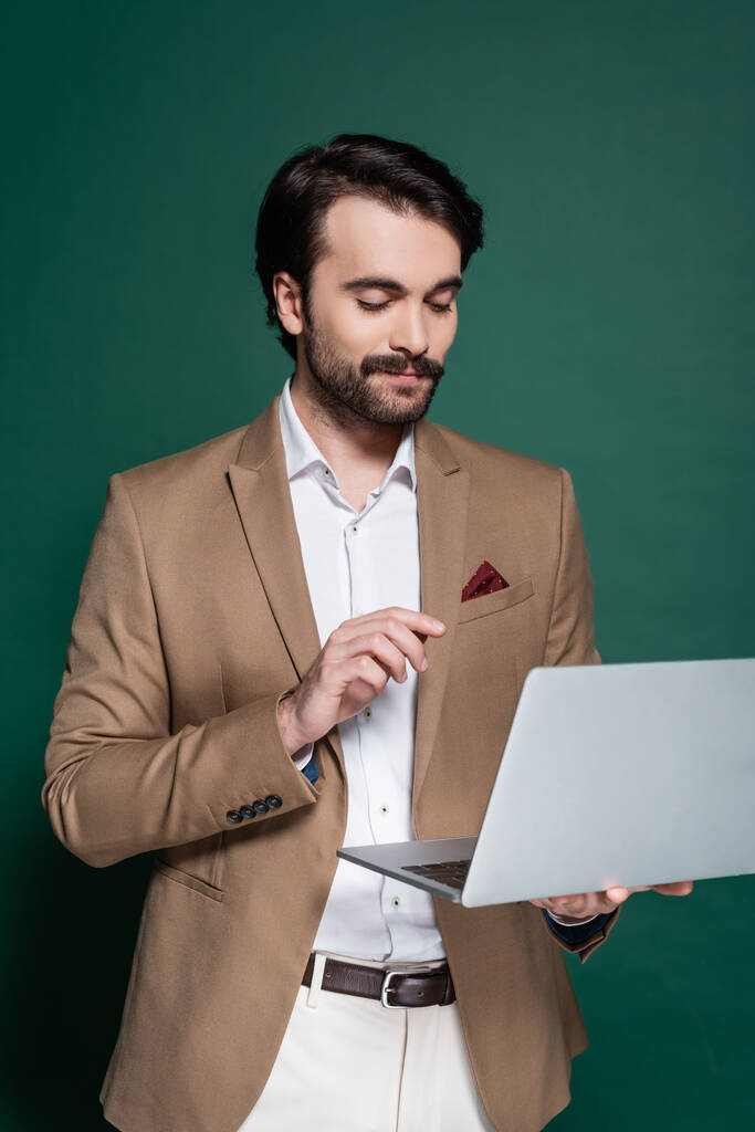 heureux jeune homme avec moustache en utilisant un ordinateur portable sur vert foncé  - Photo, image