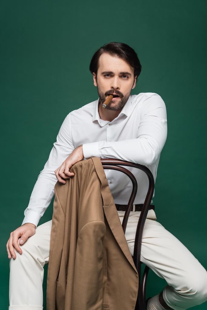 man with mustache smoking cigar and sitting on wooden chair on dark green  - Photo, Image