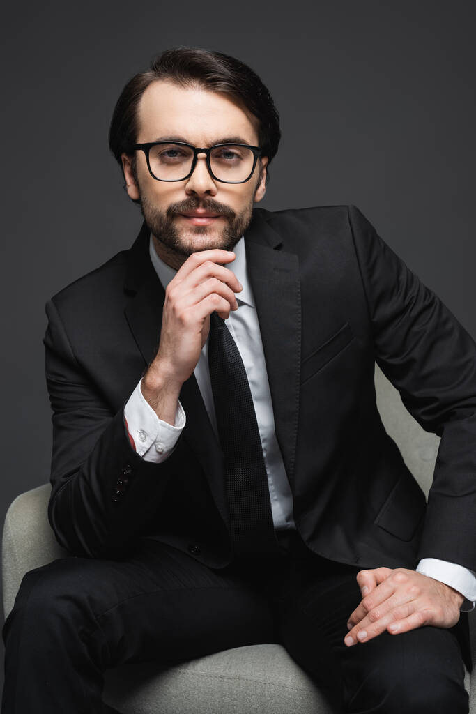 retrato de empresário de terno e óculos sentado na poltrona e olhando para a câmera no cinza escuro - Foto, Imagem