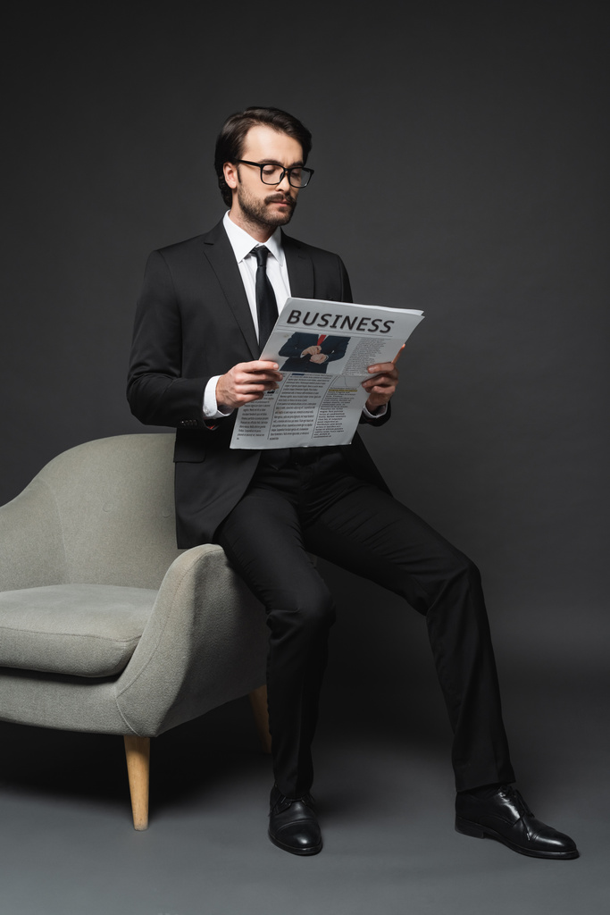 full length of businessman in suit and glasses leaning on armchair and reading business newspaper on dark grey - Photo, Image