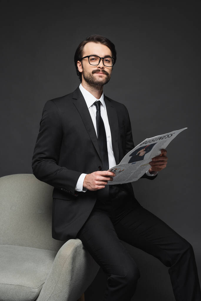 επιχειρηματίας με κοστούμι και γυαλιά ακουμπά στην πολυθρόνα και κρατά επαγγελματική εφημερίδα σε σκούρο γκρι - Φωτογραφία, εικόνα
