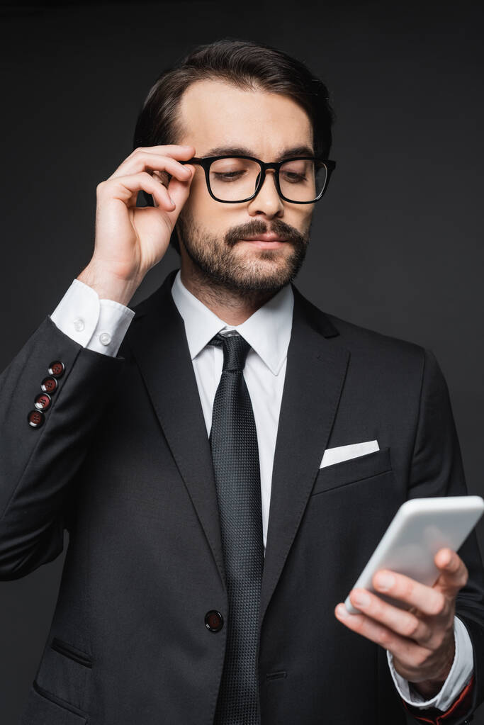 homem de negócios de terno e óculos olhando para smartphone em cinza escuro - Foto, Imagem