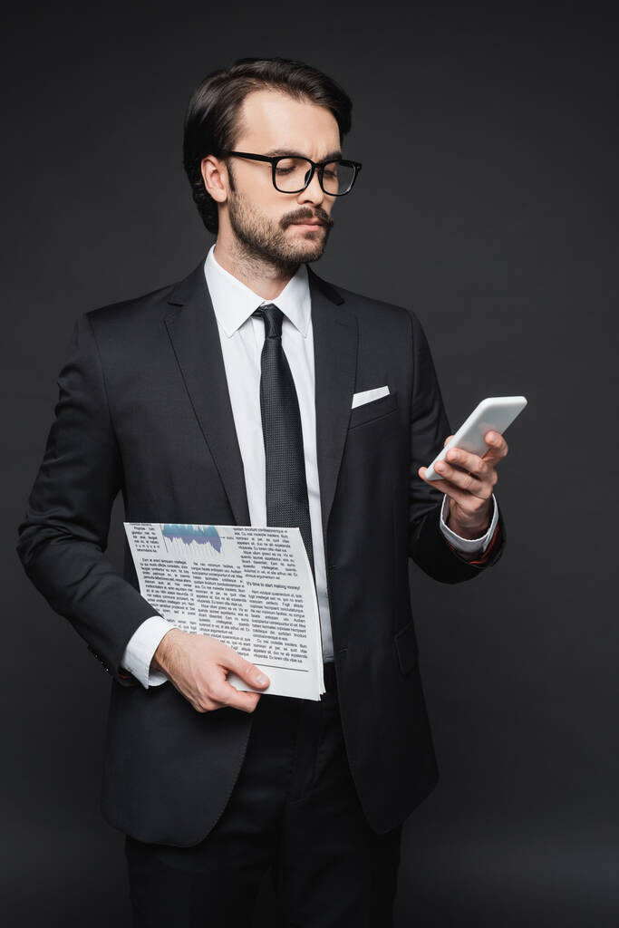 επιχειρηματίας με κοστούμι και γυαλιά κρατώντας εφημερίδα και χρησιμοποιώντας smartphone σε σκούρο γκρι - Φωτογραφία, εικόνα