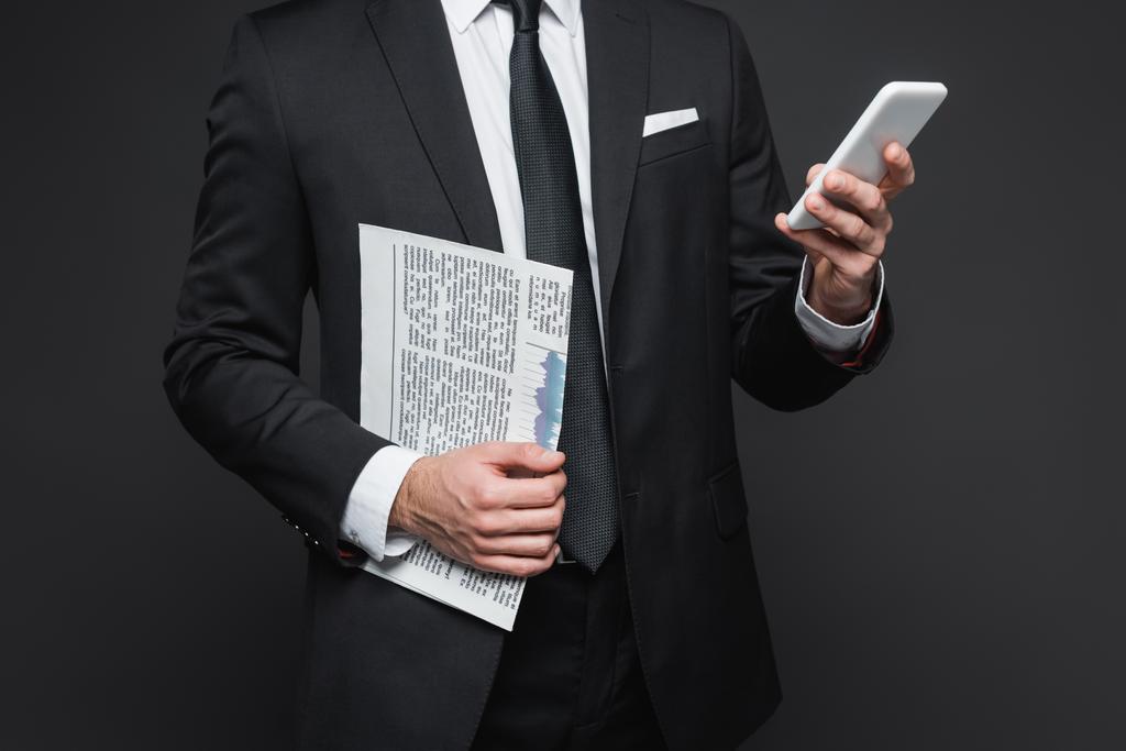 vista cortada de empresário em terno segurando jornal e usando smartphone em cinza escuro - Foto, Imagem