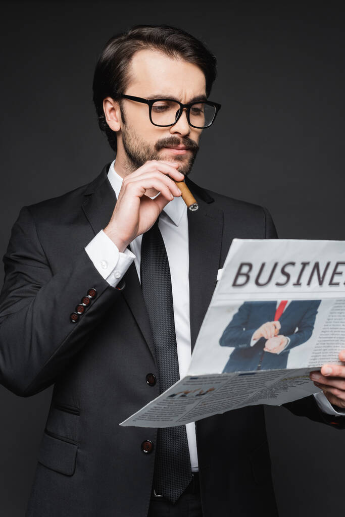 muž v obleku a brýle čtení obchodních novin a drží doutník na tmavě šedé - Fotografie, Obrázek
