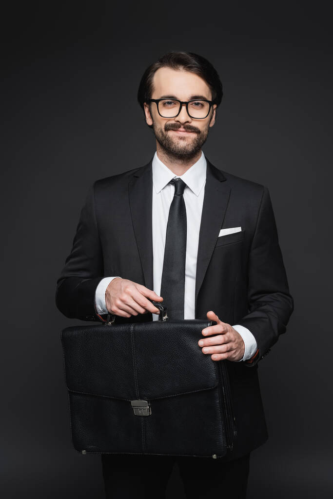homme d'affaires souriant en costume et lunettes tenant une mallette en cuir sur gris foncé - Photo, image