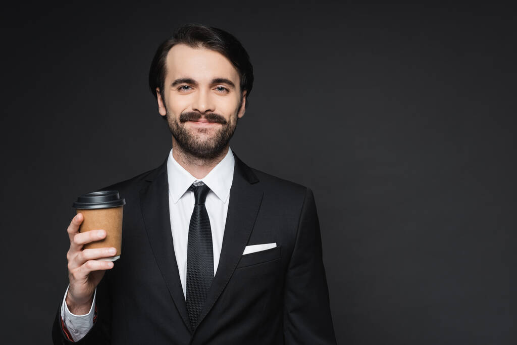 Χαρούμενος επιχειρηματίας με μουστάκι κρατώντας χάρτινο κύπελλο στο σκούρο γκρι - Φωτογραφία, εικόνα
