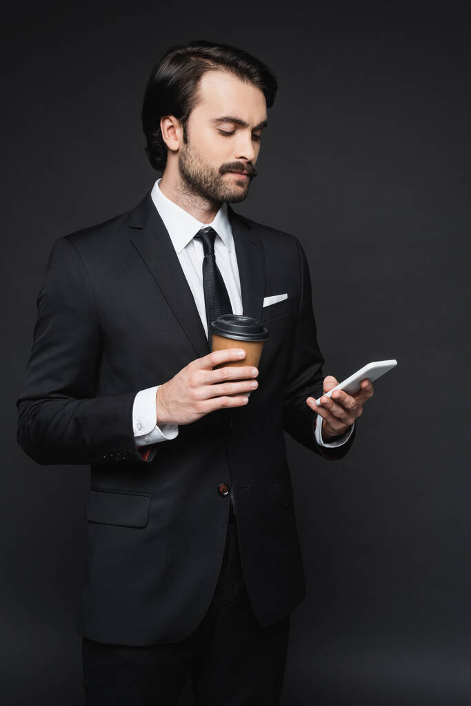 Geschäftsmann mit Schnurrbart hält Pappbecher und Smartphone auf dunkelgrau - Foto, Bild