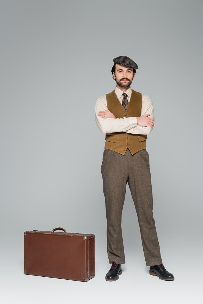 comprimento total do homem em roupas de estilo retro de pé com braços cruzados perto de saco de viagem em cinza  - Foto, Imagem