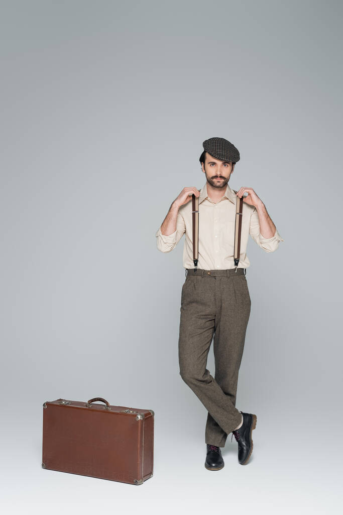 В полный рост мужчина в одежде в стиле ретро и шляпе стоит со скрещенными ногами возле дорожной сумки на сером фоне  - Фото, изображение