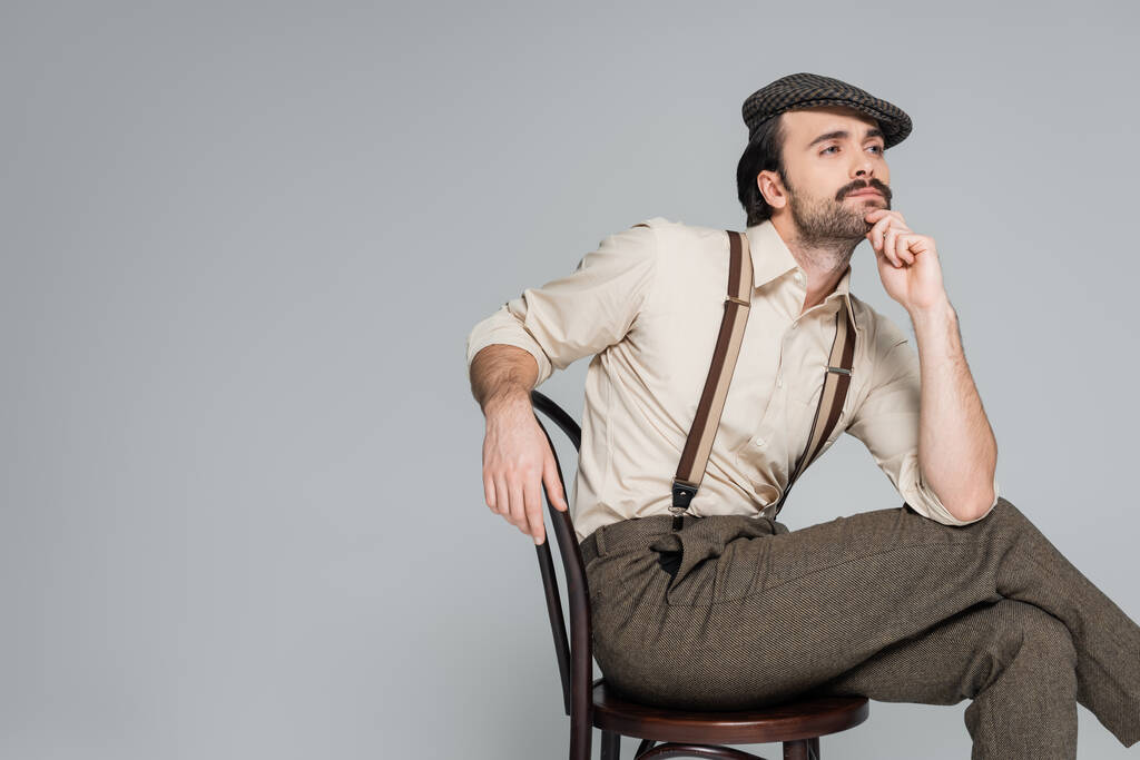 задумчивый человек с усами в ретро стиле одежды и шляпы сидя на деревянном кресле изолированы на сером  - Фото, изображение