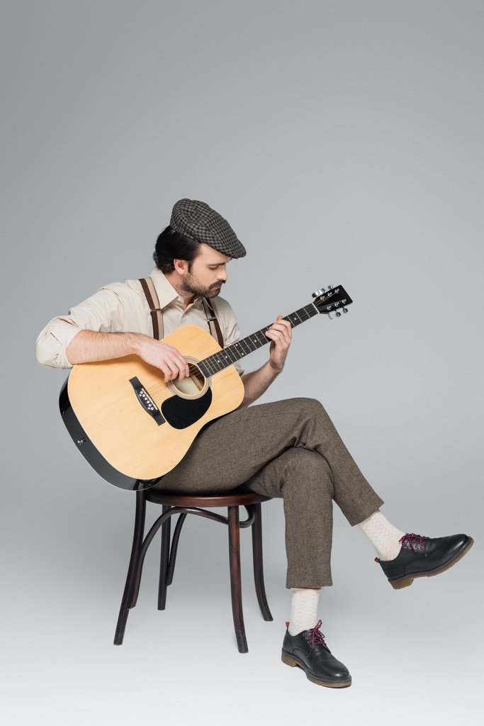 volle Länge des Mannes mit Schnurrbart im Retro-Stil Kleidung und Hut spielt Akustikgitarre auf grau  - Foto, Bild