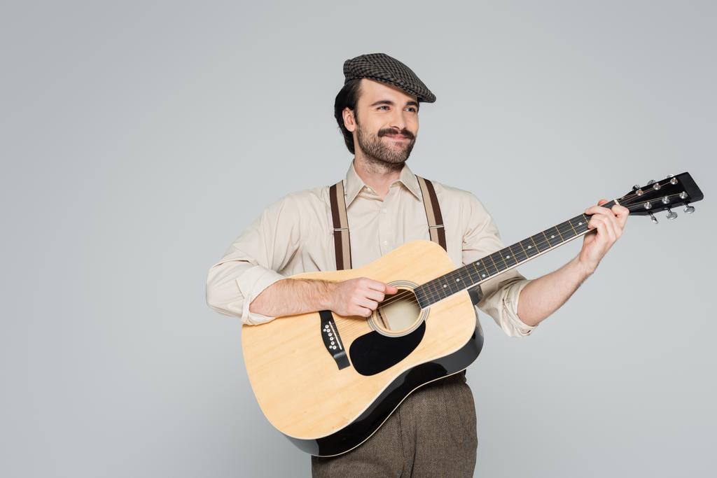 Lächelnder Mann mit Schnurrbart in Retro-Kleidung und Hut spielt Akustikgitarre isoliert auf grau  - Foto, Bild