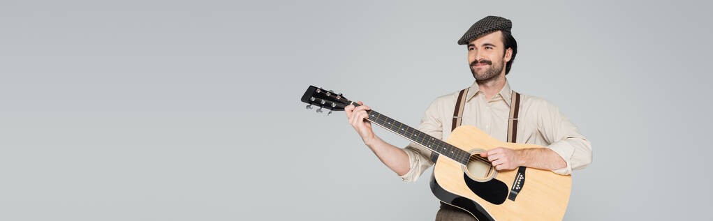 hombre sonriente con bigote en ropa de estilo retro y sombrero tocando guitarra acústica aislado en gris, bandera - Foto, Imagen